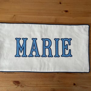 May include: A rectangular white pillow with the name "MARIE" embroidered in light blue, outlined in a darker blue. The pillow has a dark blue border and is displayed on a wooden surface.