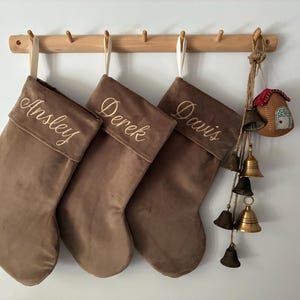 Può includere: Tre calze di Natale in velluto marrone con i nomi ricamati: Ansley, Derek e Davis. Le calze sono appese a un attaccapanni di legno. Un piccolo ornamento a forma di casa e diverse campane sono sospese a destra.