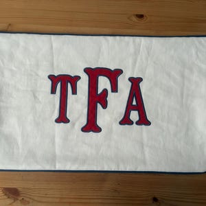 May include: White rectangular pillow with the letters "TFA" in red with a dark blue outline. The pillow has a dark blue border and is made of a textured fabric. The pillow is likely for home decor.