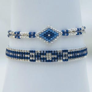 Op de afbeelding: Twee kralenarmbanden in een kleurenpalet van metallic zilver, koningsblauw en lichtblauw. De bovenste armband heeft een centraal ruitvormig motief met dubbele strengen aan de zijkanten. De onderste armband is een platte band met een patroon.