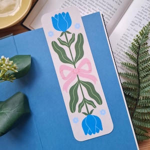May include: A bookmark with a floral design. It features blue tulips, green leaves, and a pink bow. The bookmark is placed on a blue book, with open pages visible in the background.