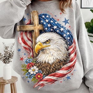 May include: A gray sweatshirt featuring a detailed graphic of an American bald eagle with a wooden cross, surrounded by the American flag and floral accents. The design incorporates red, white, and blue colors, and star details.
