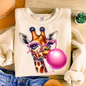 May include: Cream-colored sweatshirt featuring a giraffe wearing pink glasses and blowing a pink bubble. The giraffe has a detailed, colorful design. The sweatshirt is a casual style, suitable for everyday wear.