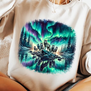 May include: A cream-coloured sweatshirt with a colourful graphic of wolves under the Northern Lights. The design shows a forest and a lake reflecting the aurora borealis, with green, purple, and blue hues.