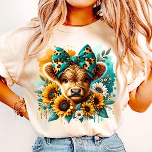 Può includere: Una maglietta bianca con una grafica di una mucca Highland marrone e bianca con un fiocco di colore turchese e stampa leopardata. La mucca è circondata da girasoli e altri fiori.