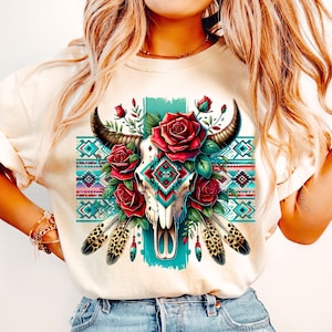 May include: Cream-coloured t-shirt featuring a detailed graphic of a bull skull adorned with red roses, feathers, and a turquoise and patterned design. The design incorporates elements of Southwestern and bohemian styles, creating a unique and artistic aesthetic.