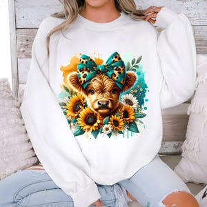 May include: White sweatshirt with a graphic of a brown and white Highland cow wearing a teal and leopard print bow. The cow is surrounded by yellow sunflowers and green leaves.