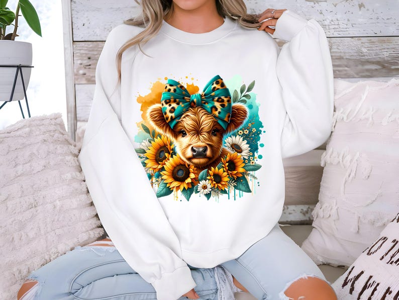 May include: White sweatshirt with a graphic of a brown and white Highland cow wearing a teal and leopard print bow. The cow is surrounded by yellow sunflowers and green leaves.