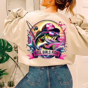 May include: A beige sweatshirt with a graphic print of a bass fish wearing a hat and sunglasses. The graphic also includes a fishing rod and the text "Reel Girls Fish".