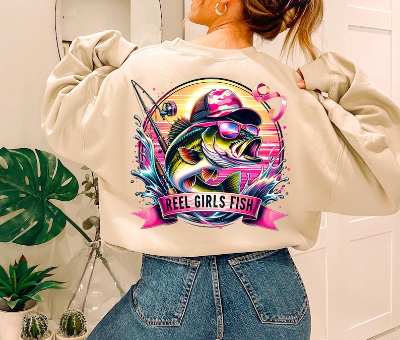 May include: A beige sweatshirt with a graphic print of a bass fish wearing a hat and sunglasses. The graphic also includes a fishing rod and the text "Reel Girls Fish".