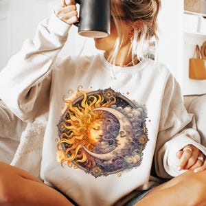 May include: A white sweatshirt with a graphic print of a sun and moon with faces. The sun is yellow and orange, and the moon is white and blue. The graphic is surrounded by stars and clouds.