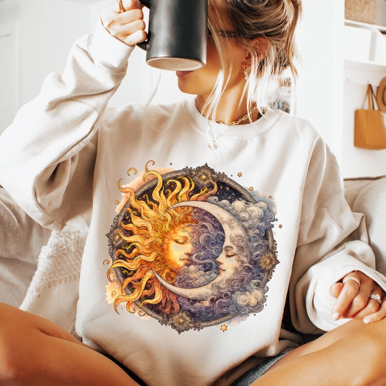 May include: A white sweatshirt with a graphic print of a sun and moon with faces. The sun is yellow and orange, and the moon is white and blue. The graphic is surrounded by stars and clouds.
