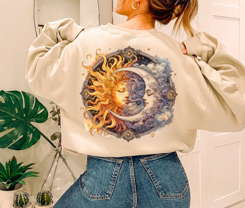 May include: A cream-colored sweatshirt with a graphic print of a sun and moon with a detailed design. The sun and moon are depicted as faces with a celestial background.