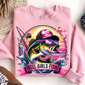 May include: Pink sweatshirt with a graphic of a bass fish wearing sunglasses and a camouflage hat. The fish is surrounded by water and a fishing rod. The text "Reel Girls Fish" is printed on a pink ribbon.