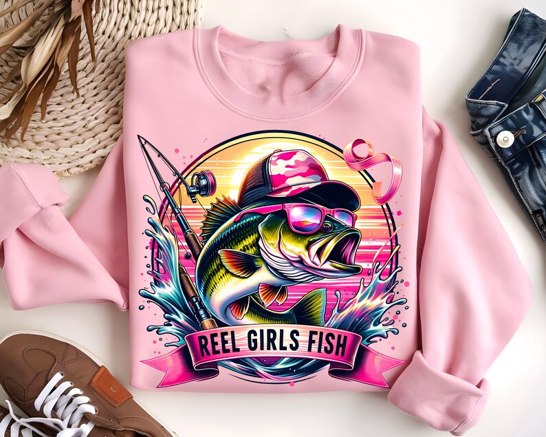 May include: Pink sweatshirt with a graphic of a bass fish wearing sunglasses and a camouflage hat. The fish is surrounded by water and a fishing rod. The text "Reel Girls Fish" is printed on a pink ribbon.
