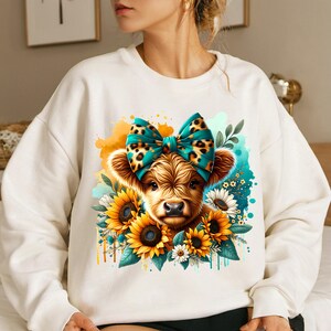 May include: White sweatshirt with a cute cartoon highland cow wearing a turquoise and leopard print bow. The cow is surrounded by sunflowers and other flowers.