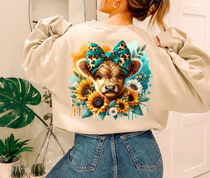 May include: A beige sweatshirt with a graphic of a brown and white Highland cow with a blue and teal bow. The cow is surrounded by sunflowers and other flowers.