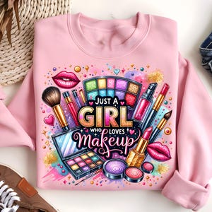 May include: Pink sweatshirt with a colorful graphic design featuring makeup brushes, eyeshadow palettes, lipstick, and the text "Just a Girl Who Loves Makeup".