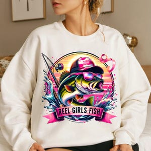 May include: White sweatshirt with a pink, blue, and green graphic of a fish wearing sunglasses and a pink hat. The graphic includes a fishing rod and the text "REEL GIRLS FISH".