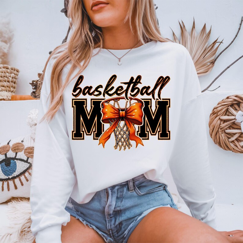 May include: White long-sleeve sweatshirt with the text "basketball mom" in black with an orange bow and basketball hoop graphic.