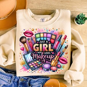 May include: A cream-colored sweatshirt with a colorful graphic design featuring makeup items, including lipstick, eyeshadow palettes, and brushes. The text "Just a Girl Who Loves Makeup" is printed in pink, purple, and yellow letters.