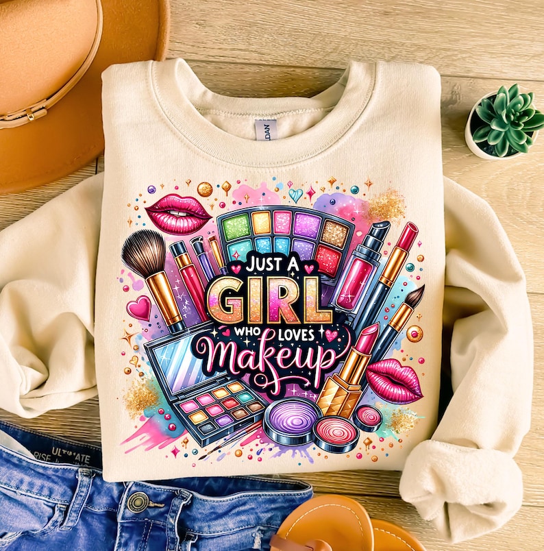 May include: A cream-colored sweatshirt with a colorful graphic design featuring makeup items, including lipstick, eyeshadow palettes, and brushes. The text "Just a Girl Who Loves Makeup" is printed in pink, purple, and yellow letters.