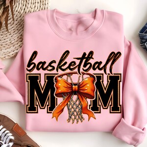 May include: Pink sweatshirt with the text "basketball mom" in black letters and a basketball hoop with an orange bow on it.