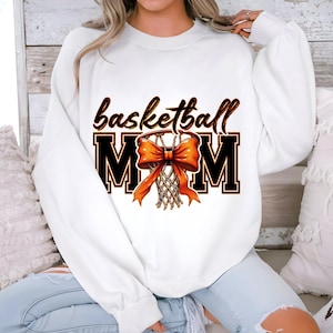 May include: White sweatshirt with the text "basketball mom" in black and orange. A basketball hoop with an orange bow is in the center of the text.