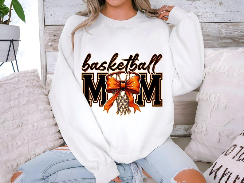 May include: White sweatshirt with the text "basketball mom" in black and orange. A basketball hoop with an orange bow is in the center of the text.