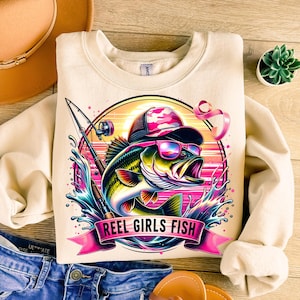 May include: A cream-colored sweatshirt with a graphic of a bass fish jumping out of the water. The fish is wearing sunglasses and a pink and camouflage baseball cap. The graphic also includes a fishing rod, a pink ribbon, and the text "Reel Girls Fish".