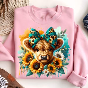 May include: Pink sweatshirt with a cute illustration of a Highland cow wearing a teal and leopard print bow. The cow is surrounded by yellow sunflowers and green leaves.