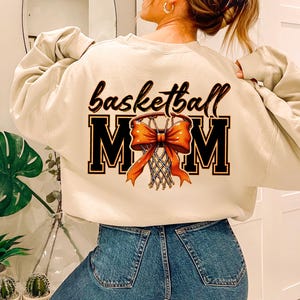 May include: A beige sweatshirt with the text "basketball mom" and a graphic of a basketball hoop with an orange bow.