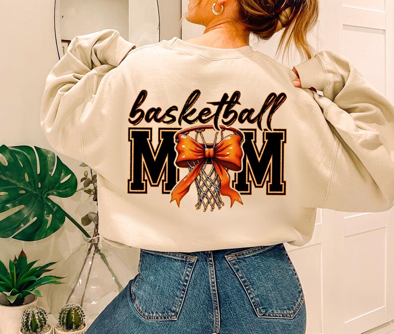 May include: A beige sweatshirt with the text "basketball mom" and a graphic of a basketball hoop with an orange bow.