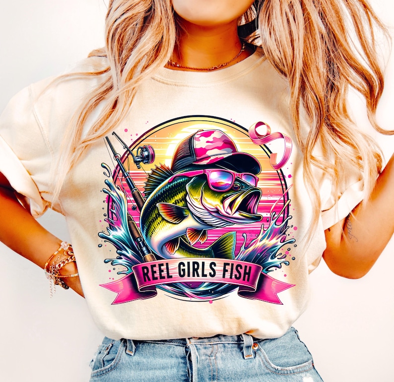May include: A white t-shirt with a pink, green, and blue graphic of a bass fish wearing sunglasses and a camouflage hat. The graphic also includes a fishing rod and the text "REEL GIRLS FISH".