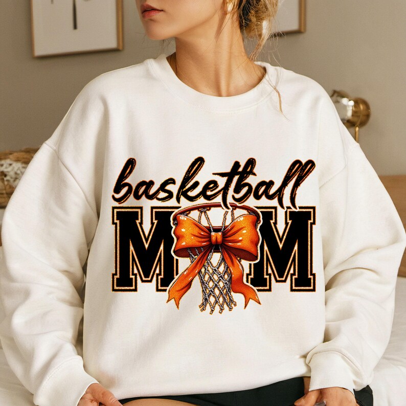 May include: White sweatshirt with the text "basketball MOM" in black with an orange basketball hoop and bow in the center.