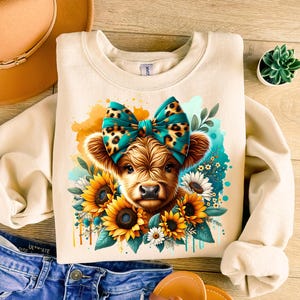 May include: A beige sweatshirt with a cute illustration of a Highland cow wearing a turquoise and brown bow. The cow is surrounded by sunflowers and other flowers.