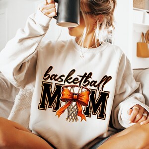 May include: White sweatshirt with the text "basketball mom" in black with an orange bow and basketball net graphic.