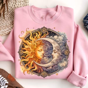 May include: Pink sweatshirt with a graphic print of a sun and moon with a celestial design. The sun and moon are depicted as faces with closed eyes.