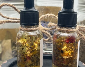 May include: Two small glass bottles with dropper tops filled with a clear liquid containing dried herbs and gold flakes. The bottles are tied with twine and are sitting on a black surface.