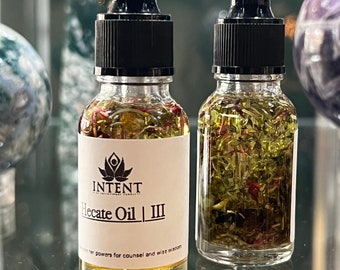 May include: Two glass bottles with dropper tops filled with a clear liquid and dried herbs. The bottle on the left has a white label with the text "INTENT" and "Hecate Oil | III" and the text "...her powers for counsel and wise wisdom".