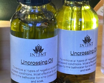 May include: Two amber glass bottles with black caps and white labels. The labels have a green logo and the text "INTENT" and "Uncrossing Oil". The text on the labels describes the oil's use to remove negative curses and crossed conditions.