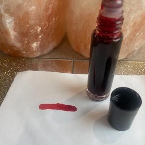 Könnte beinhalten: Eine kleine, transparente Glas-Rollerflasche gefüllt mit einer dunkelroten Flüssigkeit, neben einer schwarzen Kappe und einem roten Strich auf weißem Papier. Die Flasche hat einen roten Rollballaufsatz. Zwei große, rosa-orangefarbene Salzkristalllampen im Hintergrund.