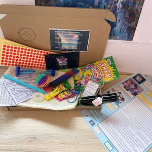 Op de afbeelding: Een bruine kartonnen doos gevuld met verschillende items, waaronder een klein zakje met kleurrijke fidget-speelgoed, een flesje etherische olie, een klein zakje snoep, een klein zandloper en een vel papier met instructies. De doos is gelabeld met de tekst "Self Help Therapy" en bevat social media-handvatten voor de verkoper.