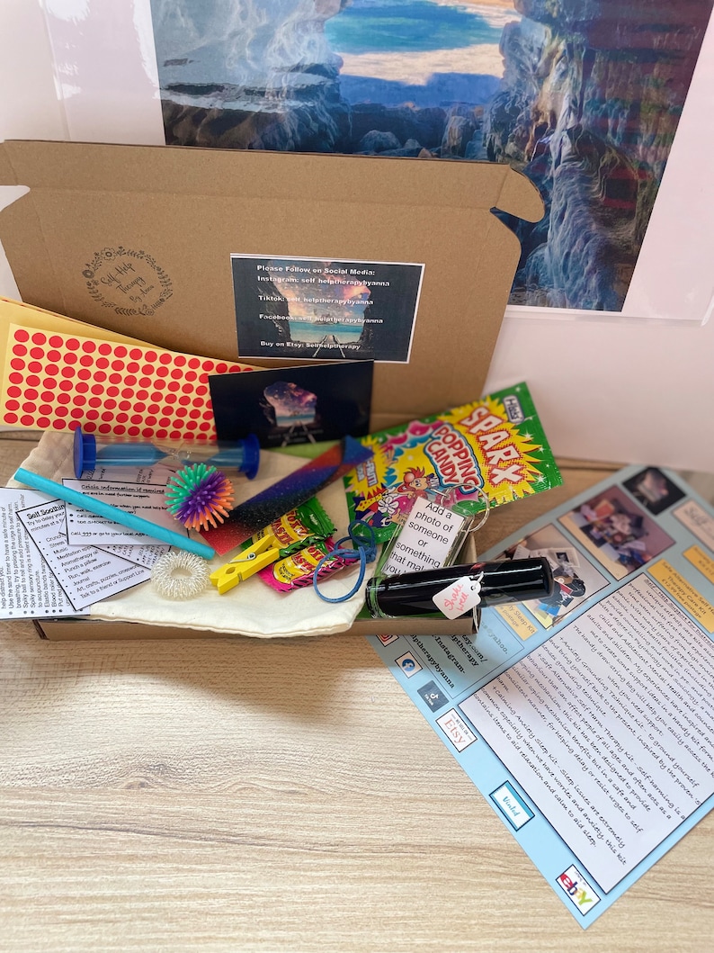 Op de afbeelding: Een bruine kartonnen doos gevuld met verschillende items, waaronder een klein zakje met kleurrijke fidget-speelgoed, een flesje etherische olie, een klein zakje snoep, een klein zandloper en een vel papier met instructies. De doos is gelabeld met de tekst "Self Help Therapy" en bevat social media-handvatten voor de verkoper.
