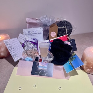 Könnte beinhalten: Eine hellgelbe Geschenkbox mit einer rosa-weißen Seidenpapierauskleidung. Die Box enthält eine Vielzahl von Artikeln, darunter eine Lavendel-Augenmaske, eine kleine Flasche Flüssigkeit, eine schwarze Augenmaske, ein kleines Notizbuch, eine schwarze, flauschige Socke, eine kleine Tüte Kristalle, eine kleine Flasche Nagellack, eine Postkarte mit einer Strandszene und eine Visitenkarte mit der Aufschrift "Self-Help Therapy by Anna".