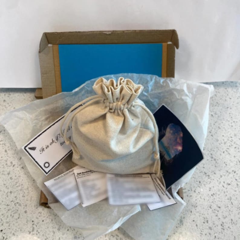 K&ouml;nnte beinhalten: Ein kleiner, beiger Stoffbeutel mit Kordelzug befindet sich in einem Karton mit blauer Papierauskleidung. Die Schachtel enth&auml;lt auch eine wei&szlig;e Karte mit dem Text "Es ist in Ordnung, wenn du dich heute nicht zu gl&uuml;cklich f&uuml;hlst." und ein kleines, gedrucktes Bild einer Galaxie.