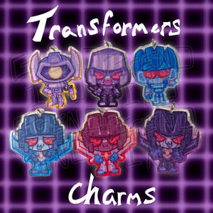 May include: A collection of six glittery Transformers charms in various colors, including purple, blue, and red. The word "Transformers" is written in white script at the top, and "Charms" is at the bottom.