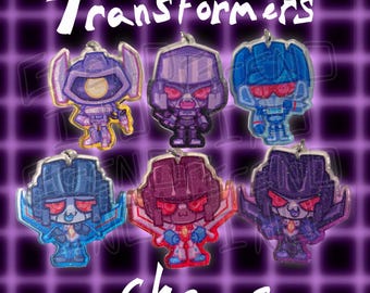 Op de afbeelding: Een verzameling van zes glitterende Transformers-bedels in verschillende kleuren, waaronder paars, blauw en rood. Het woord "Transformers" staat in witte letters bovenaan en "Charms" onderaan.