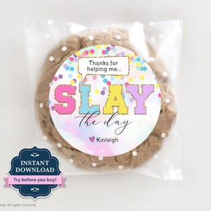Può includere: Un grande biscotto con gocce di cioccolato in un sacchetto di cellophane trasparente con pois bianchi. Il biscotto ha un adesivo circolare con la scritta "SLAY the day" in lettere colorate, con il nome "Kinleigh" sotto. L'adesivo dice anche "Grazie per avermi aiutato..."
