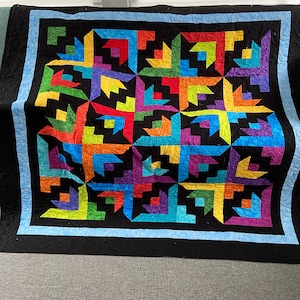 May include: A rectangular quilt featuring a repeating geometric pattern of interlocking shapes in vibrant colors including red, orange, yellow, green, blue, and purple. The design is set against a black background with a light blue border.